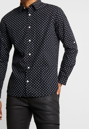 Homme portant une chemise noire à manches longues avec un motif en losanges blancs et un pantalon noir, main dans la poche.