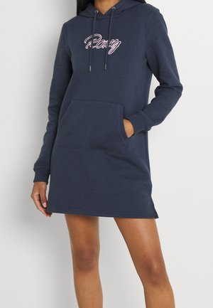 Femme portant une robe sweat à capuche bleu marine avec une poche avant et le logo « Roxy » en script rose sur la poitrine.