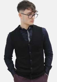 Zwarte gebreide vest met een V-hals ontwerp, voorzien van een knoopsluiting aan de voorkant, gedragen over een marineblauw shirt met een gestreepte kraag.