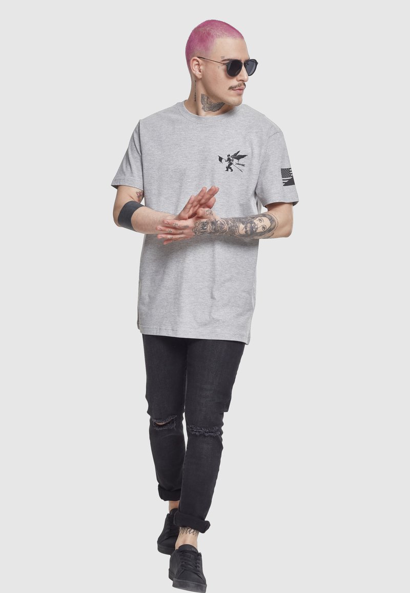 Bărbat cu păr roz și ochelari de soare, purtând un tricou gri cu imprimeu și blugi negri zdrențuiți, mergând cu mâinile împreunate, surprins în mișcare.