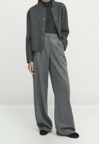 Veste en mélange de laine grise avec des boutons-pression, associée à un pantalon large taille haute gris. Le modèle se tient avec les mains dans les poches.
