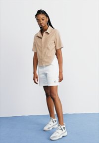 Beige korte mouwen shirt met kraag, witte shorts met een zwart logo, en grijze sportschoenen met witte accenten. Staande op een blauwe achtergrond.
