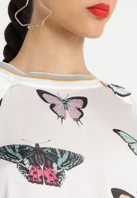 Camisa blanca con estampados de mariposas en varios colores, con un cuello redondo a rayas azules y amarillas, textura suave y mangas cortas.