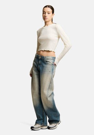 Młoda kobieta ubrana w kremowy, długi crop top, wyblakłe luźne jeansy, białe trampki i kolczyki koła, stoi z jedną ręką w kieszeni.