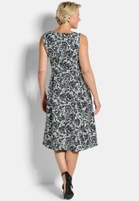 Vrouw met de rug naar de camera, gekleed in een mouwloze zwart-witte bloemenjurk en zwarte hoge hakken tegen een effen achtergrond.