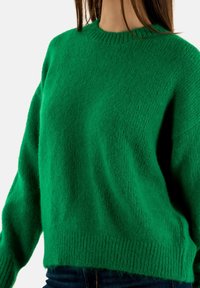 Suéter verde de punto con cuello redondo, corte holgado y puños acanalados. Textura suave y una apariencia ligeramente difusa en todo el suéter.