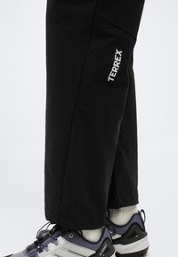 Κατώτερα άκρα που φορούν μαύρο παντελόνι TERREX και γκρι-μωβ παπούτσια πεζοπορίας με λευκές κάλτσες σε λευκό φόντο.