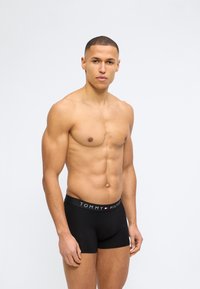 Νεαρός μυώδης άνδρας που στέκεται, φοράει μαύρα μποξεράκια Tommy Hilfiger, στραμμένος προς τα εμπρός με ουδέτερη έκφραση σε λευκό φόντο.