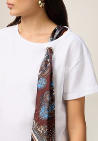 Șal din mătase de culoare maro cu modele paisley albastre, fixat cu un inel argintiu, așezat peste un tricou alb cu mânecă scurtă.