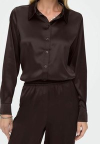 Femme portant une chemise boutonnée à manches longues en satin marron foncé rentrée dans un pantalon assorti avec une taille élastique.