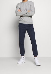 Šedá mikina s logem, navy jogger kalhoty s elastickými manžetami a bílé sportovní boty s texturovanými tkaničkami, stylizováno na jednobarevném pozadí.