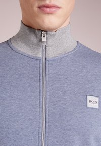 Primo piano di una persona che indossa un maglione blu con zip, colletto grigio a coste e un'etichetta con il logo visibile di Hugo Boss.