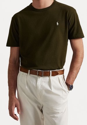 Oliwkowa koszulka bawełniana z okrągłym dekoltem i krótkimi rękawami, z małym białym logo na piersi, zestawiona z beżowymi spodniami i brązowym paskiem.