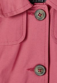 Chemise rose à manches longues, avec col, présentant des poches arrondies, un bouton décoratif à quatre trous et des détails de coutures subtils.
