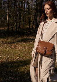 Brun læder crossover taske med et struktureret design, klaplukning og justerbar rem, parret med en lys beige trenchcoat.