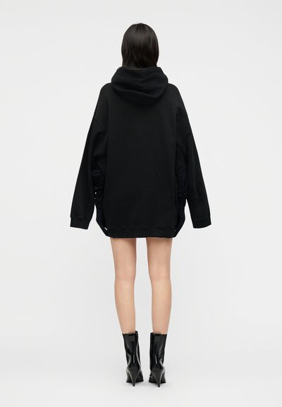 Άτομο φορώντας φαρδιά μαύρη κουκούλα, μαύρα γάντια και μαύρα μποτάκια με ψηλό τακούνι, στέκεται γυρισμένο προς τα πίσω σε απλό φόντο.