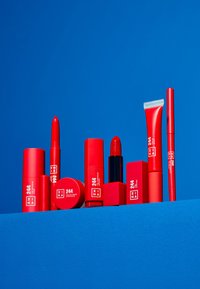 Red cosmetic products arranged on a blue background, featuring various shapes and sizes, including lipsticks, cream eyeshadow, and pencils.