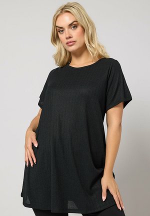 Femme blonde portant une tunique noire à manches courtes et amples, posant avec une main sur la hanche devant un fond gris uni.