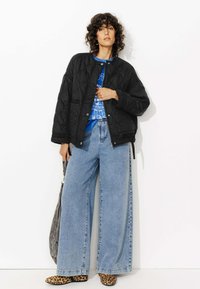 Veste bomber noire matelassée, t-shirt graphique bleu, jeans en denim léger larges, et chaussures à imprimé léopard. Sac gris porté sur une épaule.