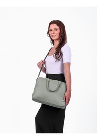 Grüne Lederhandtasche mit diagonaler Naht, zwei Griffen und einem verstellbaren Schultergurt, gehalten von einer Person in einem schwarzen Outfit.
