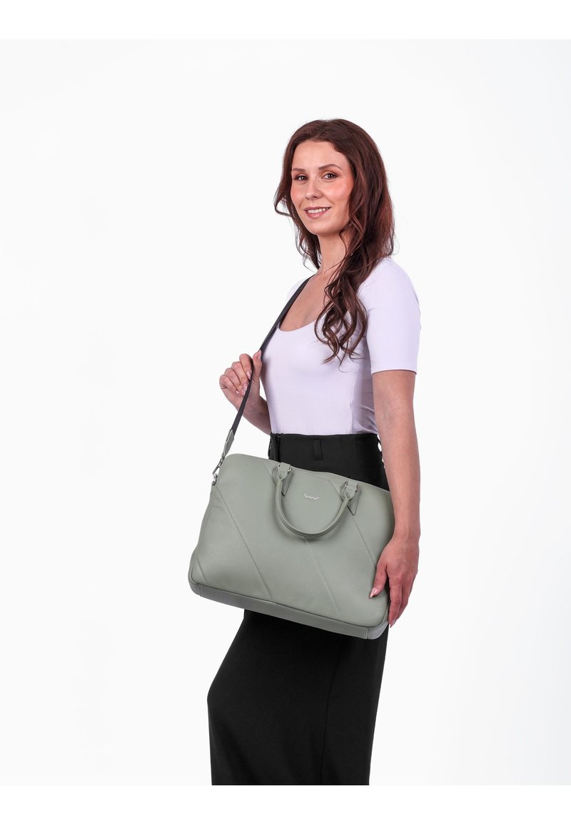 Grüne Lederhandtasche mit diagonaler Naht, zwei Griffen und einem verstellbaren Schultergurt, gehalten von einer Person in einem schwarzen Outfit.