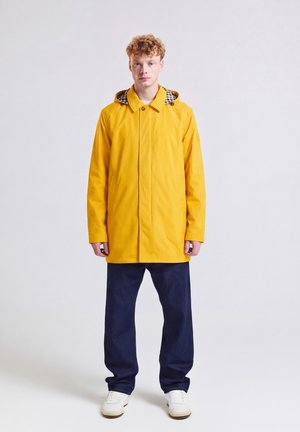 Junger Mann mit lockigem Haar steht, trägt einen gelben Kapuzenmantel, dunkelblaue Hose und weiße Sneakers vor einem einfarbigen hellen Hintergrund.