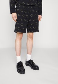 Shorts noirs avec un motif répétitif de visages souriants jaunes, fabriqués en tissu doux, associés à des chaussures en cuir noires et des chaussettes blanches.