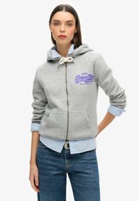 Felpa grigia con zip e cappuccio regolabile, tasche frontali e logo ricamato viola. Indossata sopra una camicia a collo azzurro, abbinata a jeans blu.
