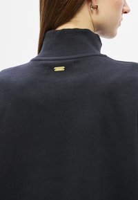 Πουλόβερ με ρούχα λαιμόκοψη και ριμπ ύφανση σε ναυτικό μπλε χρώμα, με χρυσή ετικέτα πίσω. Διαθέτει εφαρμογή που αγκαλιάζει το σώμα και μαλακή υφή υφάσματος.