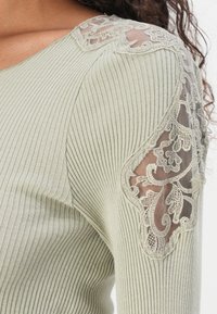 Lys grønn ribbestrikket topp med blonder på skuldrene, som gir en teksturert kontrast mot et mykt, transparent utseende.