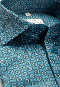 Close-up van een overhemdkraag met een blauwgroen patroon, blauwe knopen en cirkelvormige geometrische ontwerpen.