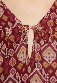Primo piano di un blusa bordeaux a motivo con scollo a V annodata con un fiocco, caratterizzata da lacci con punta metallica.