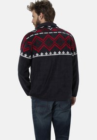 Fleece-Pullover in Marineblau mit einem bunten geometrischen Muster in Rot und Weiß. Verfügt über einen Kragen und lange Ärmel, kombiniert mit dunklen Jeans.