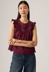 Top bordeaux senza maniche con dettagli in pizzo testurizzato, maniche a volant e orlo scallop, abbinato a jeans in denim blu.