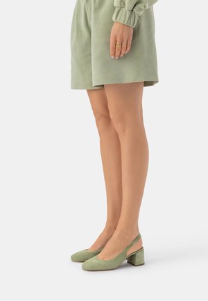 Kobieta nosząca jasnooliwkowe zamszowe sandały na słupku z paskiem z tyłu i pasujące krótkie spodenki, stojąca na jednolitym białym tle.