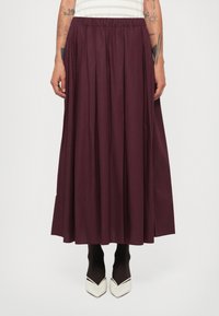 Jupe longue plissée en tissu bourgogne profond avec une taille élastique. Elle tombe en dessous du mollet et est associée à des collants noirs et des chaussures blanches à bouts pointus.