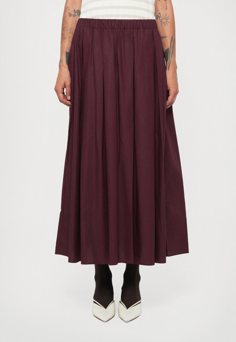 Jupe longue plissée en tissu bourgogne profond avec une taille élastique. Elle tombe en dessous du mollet et est associée à des collants noirs et des chaussures blanches à bouts pointus.