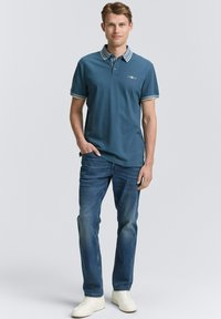 Türkises Polo-Shirt mit kontrastierendem Kragen und Ärmelbesatz, kombiniert mit blauen Jeans und weißen Sneakers. Lässiger Schnitt, minimale Logos.