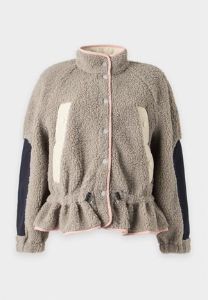 Kvinde står iført en beige tekstureret jakke med sorte albue-patches, brede beige bukser og mørkebrune snørestøvler på hvid baggrund.