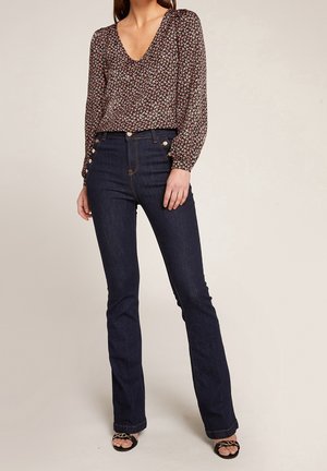 Kvinde iført mørke højhælede jeans med knapper i siden, en langærmet blomsterbluse og sorte åbne hæle med kædedetalje.