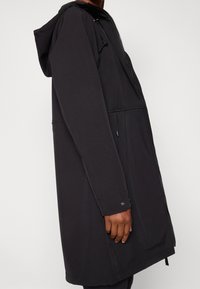 Manteau long noir avec capuche ajustée, fermeture éclair, poches latérales et texture lisse et extensible. Le design met en avant une silhouette droite.