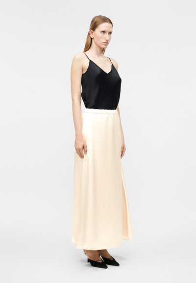 Γυναίκα που στέκεται φορώντας μαύρο αμάνικο τοπ με λεπτές τιράντες, μακριά κρεμ φούστα και μαύρα ψηλοτάκουνα παπούτσια με μυτερή μύτη μπροστά σε απλό ανοιχτό φόντο.