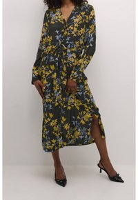 Robe maxi fleurie en vert foncé avec des motifs jaunes et bleus. Présente une taille cintrée, des manches longues et une fente haute sur la cuisse. Portée avec des escarpins noirs.