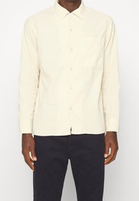 Jasnobeżowa koszula na długi rękaw na guziki z kołnierzykiem, kieszenią z przodu i gładką fakturą. Noszona na ciemnych spodniach, o luźnym kroju.