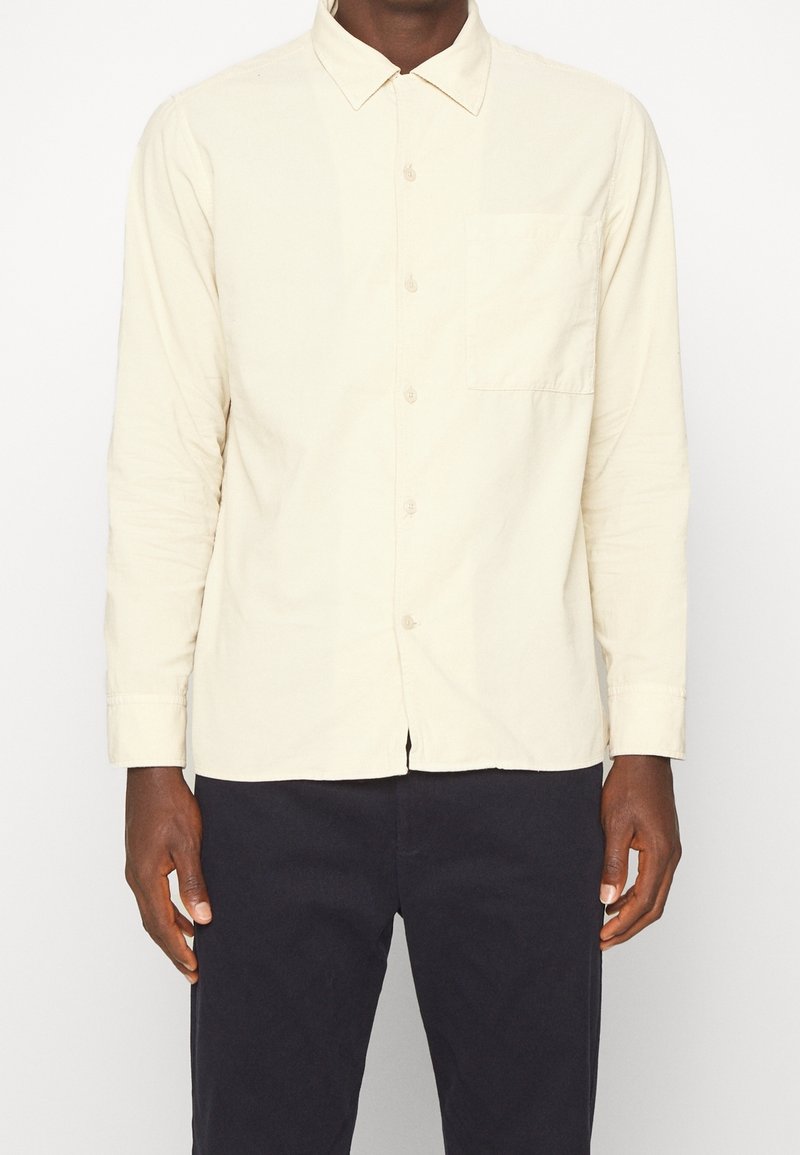 Jasnobeżowa koszula na długi rękaw na guziki z kołnierzykiem, kieszenią z przodu i gładką fakturą. Noszona na ciemnych spodniach, o luźnym kroju.
