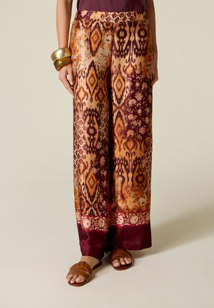 Vrouw draagt een broek met wijde pijpen met oranje, kastanjebruine en beige geometrische en bloemmotieven en bruine sandalen.
