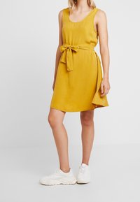 Geel mouwloos jurkje met een tailleband, gladde textuur, casual ontwerp, kniehoogte, gecombineerd met witte sneakers op een neutrale achtergrond.