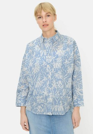 Kvinde iført en langærmet skjorte med knapper og et blåt og hvidt abstrakt bladmotiv samt en lyseblå denimnederdel.