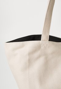Leichte beigefarbene Canvas-Tote-Tasche mit strukturierter Oberfläche, schwarzem Innenfutter und robusten Trägern zum Tragen. Der obere Rand ist genäht.