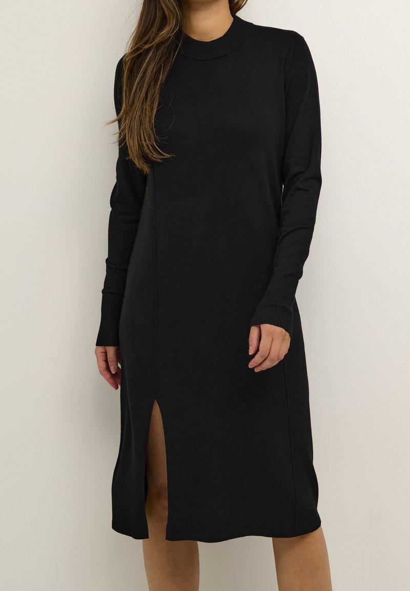 Femme portant une robe midi noire à manches longues, col rond et fente à l'avant, debout sur un fond clair uni.